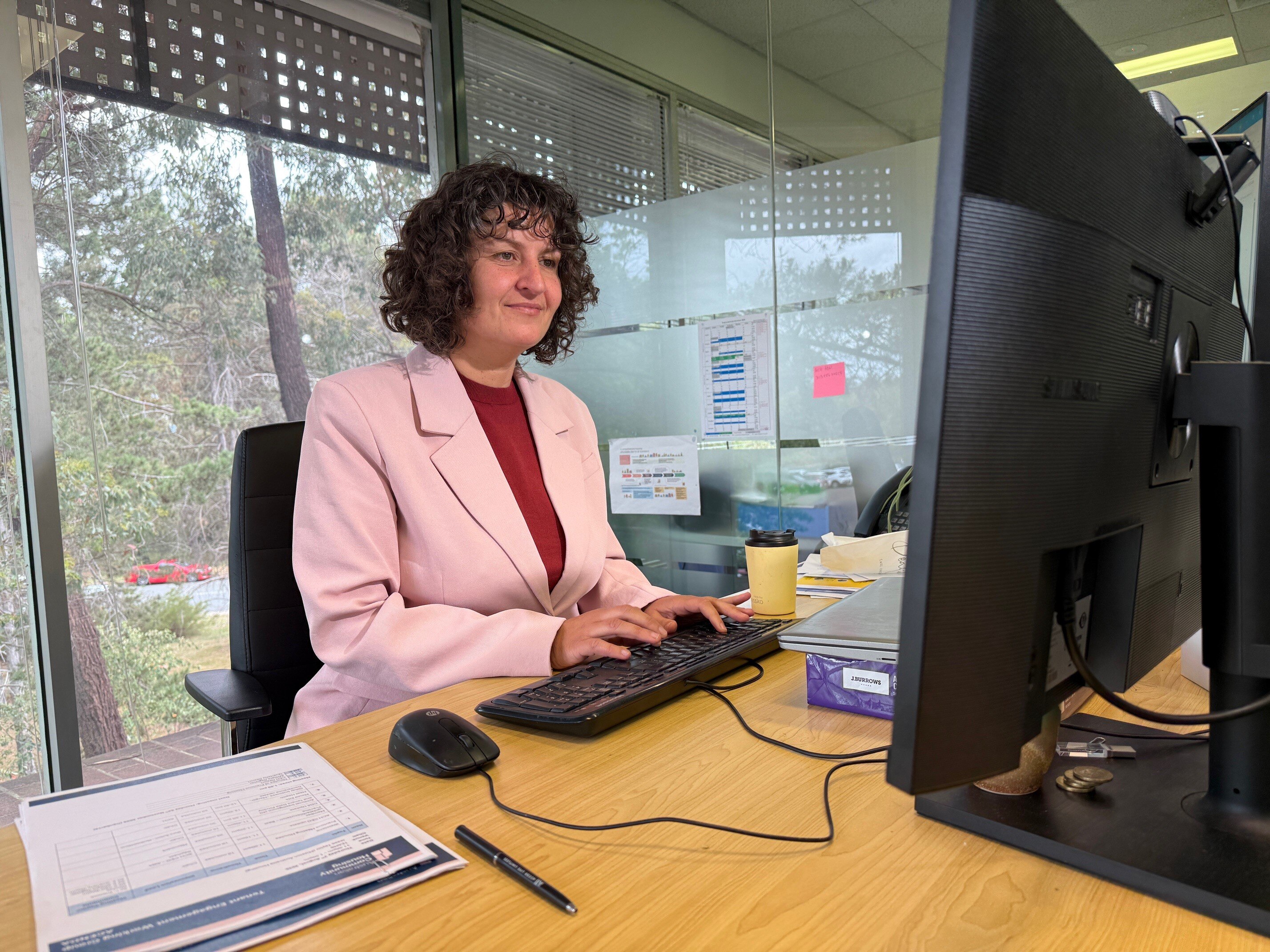 A lady wearing a pink blush blazer and red shirt sits at an office desk typing on a keyboard while looking at a computer screen.