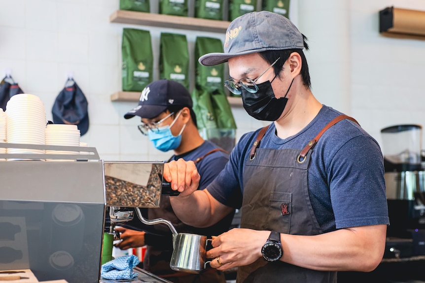 Two men wearing aprons and face masks stand at a coffee machine holding a milk jug.