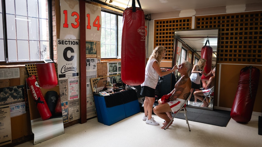 McBride sits on a folding chair in a boxing gym as a makeup artist puts make up on his face. There are punching bags nearby.