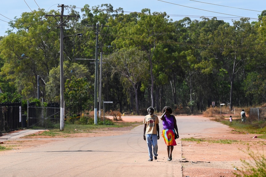 a wide shot of two people walking down an asphalt and dirt road in the community of aurukun