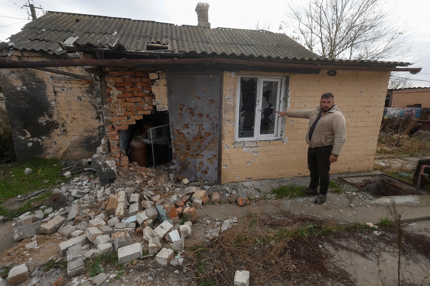 A man points to the crumbling wall of a house damaged by military strikes.