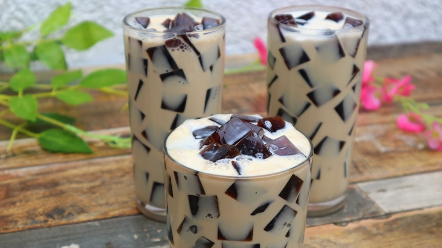 Black coloured jelly cubes in brown milk inside three glasses, set on wooden table with a plant in background out of focus.