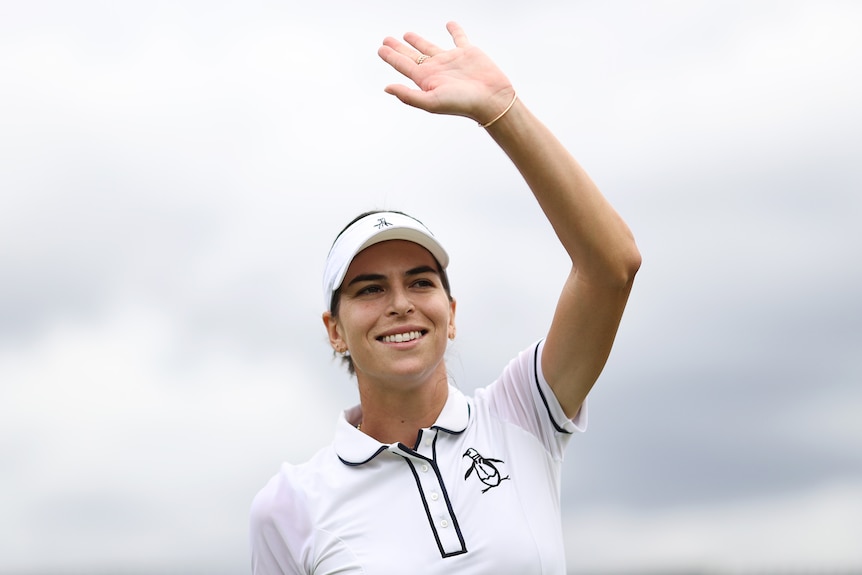 Aja Tomljanovic, wearing white tennis clothes, raises a hand to wave in front of a cloudy sky.