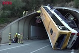 A bus is nearly upside down at the exit of a tunnel with firefighters working to stabilise it and get people out of it
