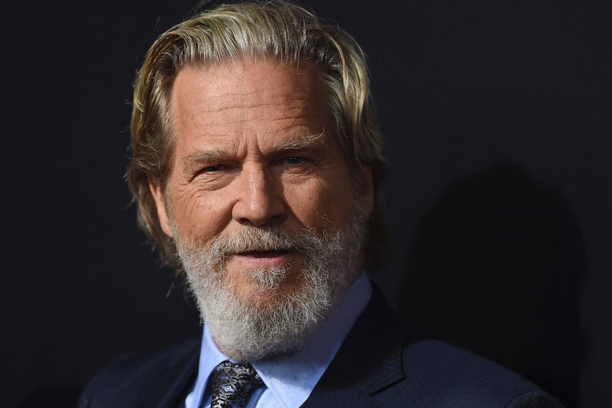 Jeff Bridges looks to the camera with a slight smile. He wears a navy suit, light blue shirt, patterned tie and has a beard.