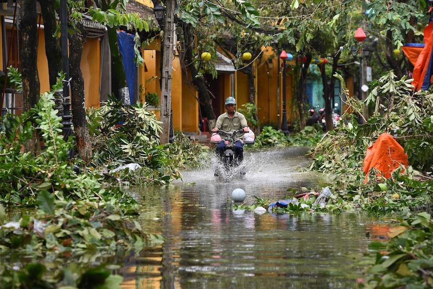 A man rides a motorbike in a flooded street through water that comes about half way up the wheel.