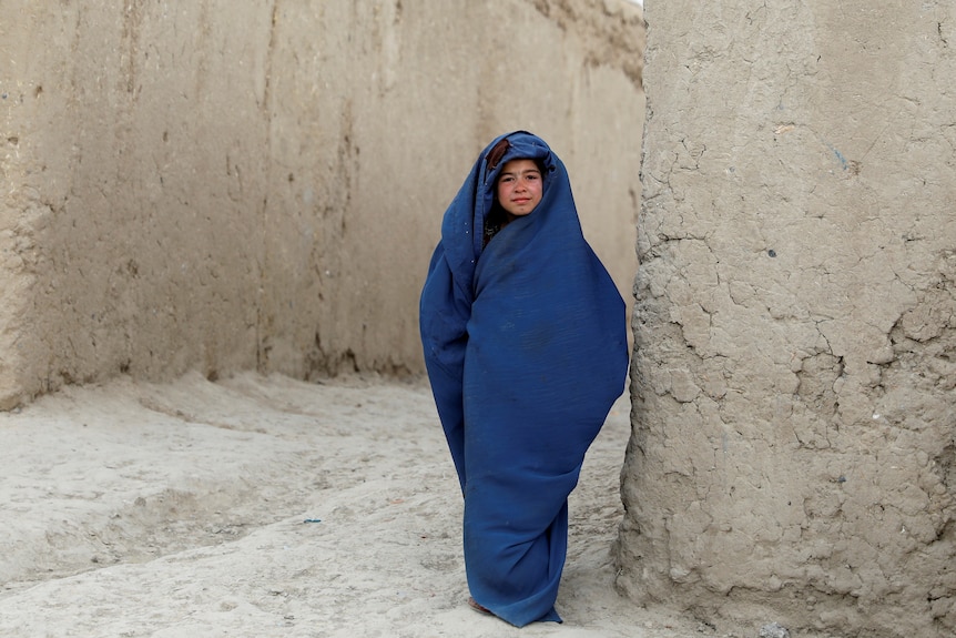 A young girl with a blue burka wrapped around her looking strong while standing next to a white wall