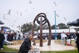 A woman releases white doves from a cage on the ground in front of a crowd gathered on a grey and rainy day.