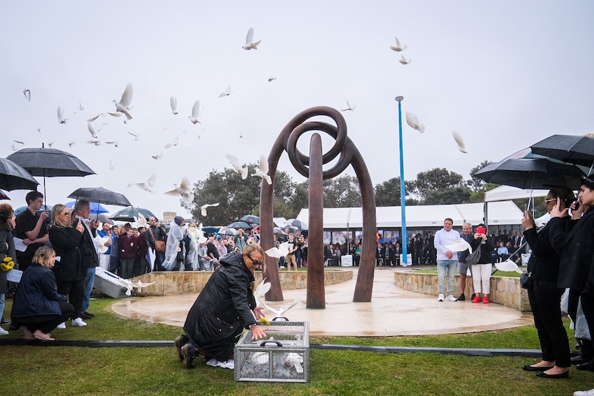 A woman releases white doves from a cage on the ground in front of a crowd gathered on a grey and rainy day.