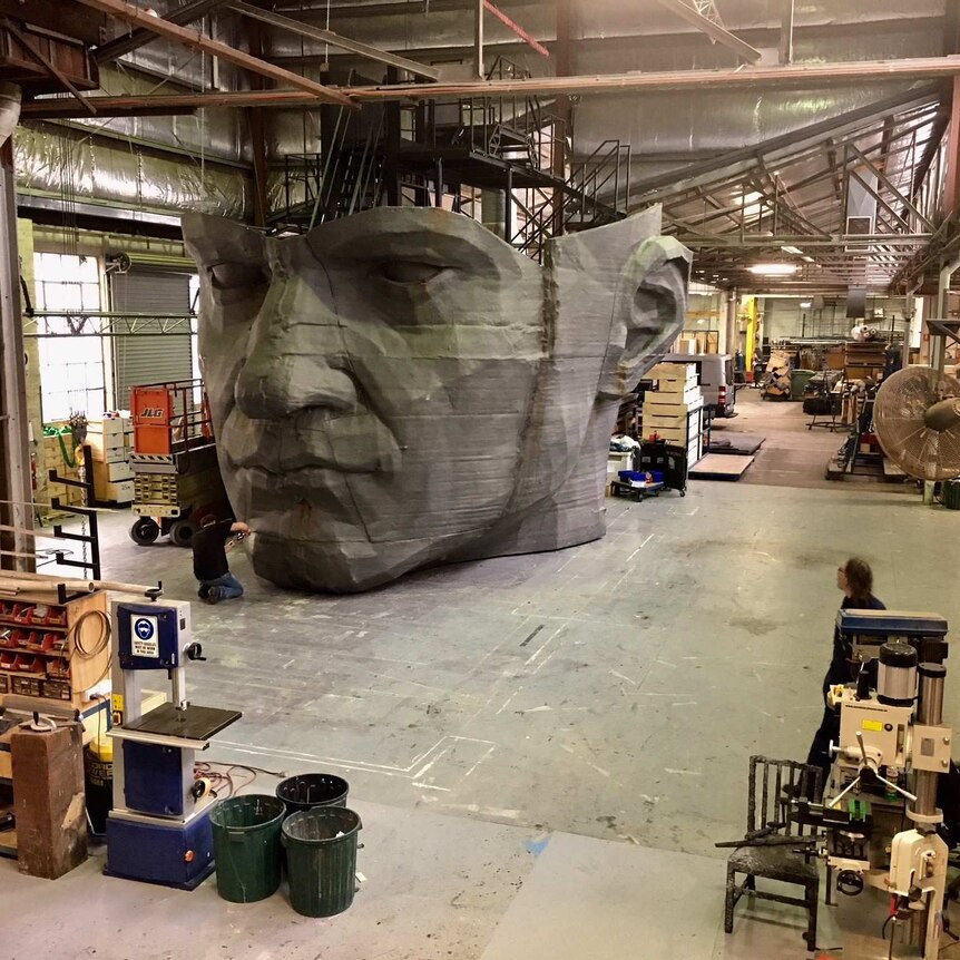 In a large industrial workshop, a man works on a giant prop of a stone head.
