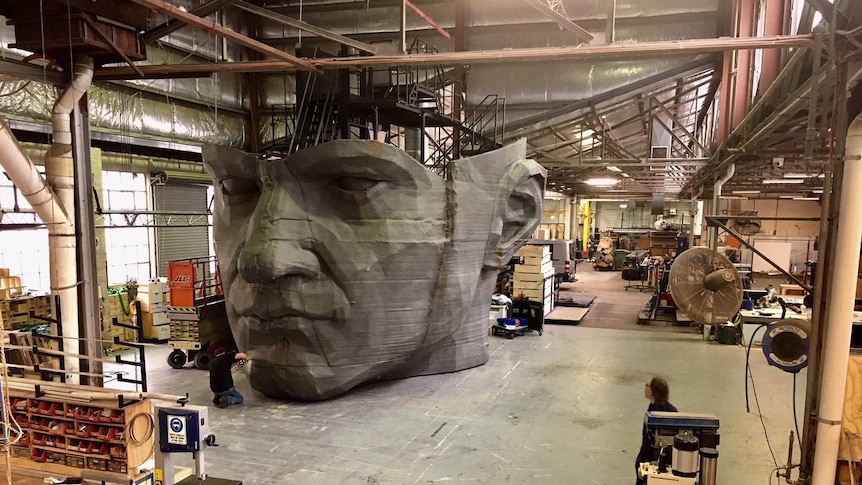In a large industrial workshop, a man works on a giant prop of a stone head.