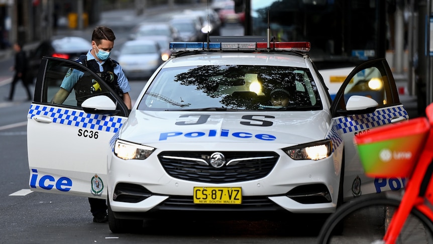 A policeman wearing a mask looks down at his patrol car with doors on either side of the vehicle open in a Sydney street