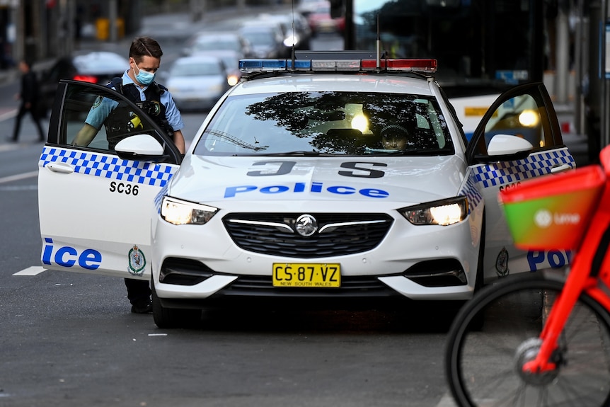 A policeman wearing a mask looks down at his patrol car with doors on either side of the vehicle open in a Sydney street