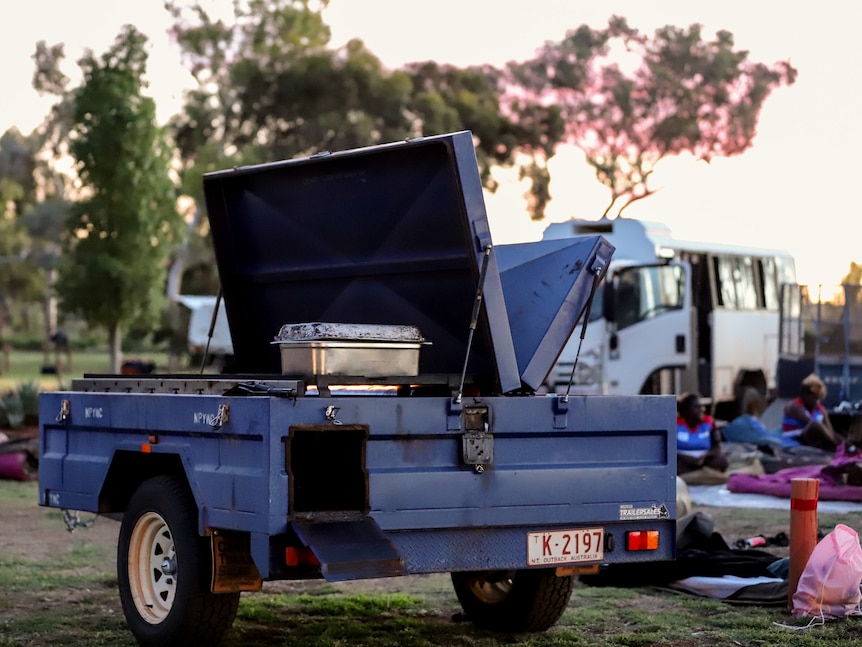 A barbecue on the back of a blue trailer sits in a campground amid camping equipment and a bus at sunset