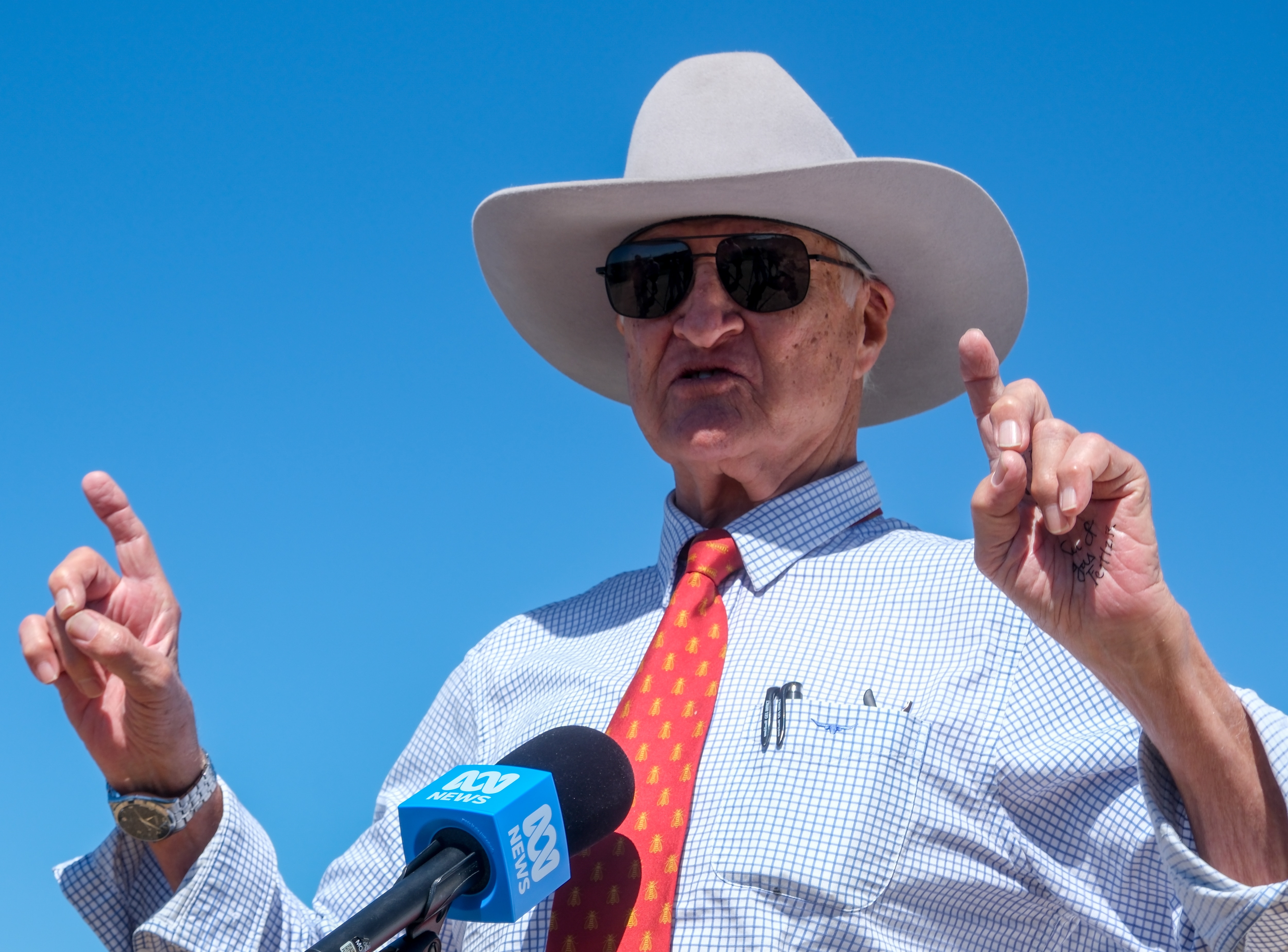 A man wearing a cowboy-style hat and a shirt and tie holds both hands up while speaking