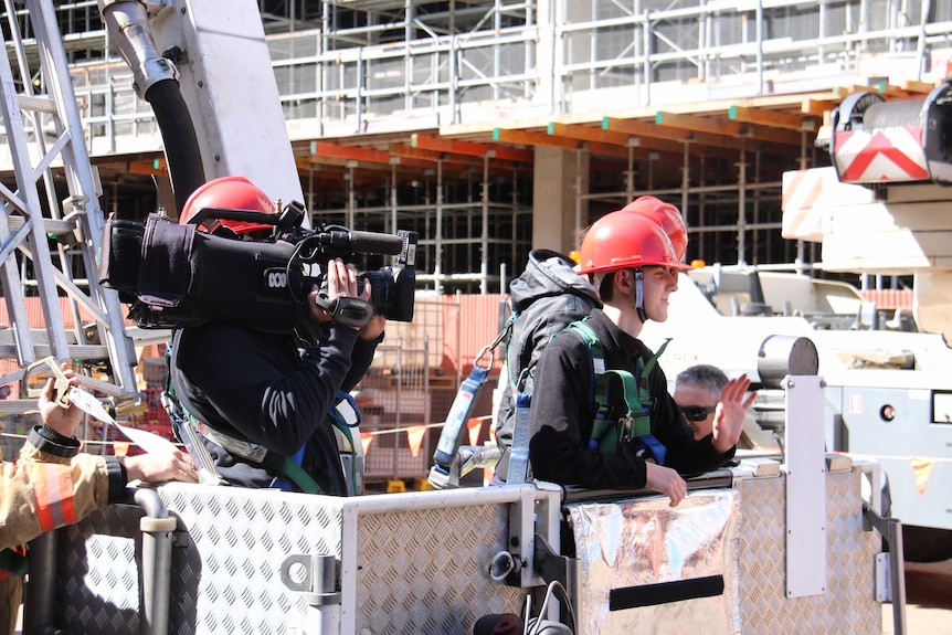 Haidarr Jones hidden behind camera filming two men wearing red firemen helmets in a cherry picker.