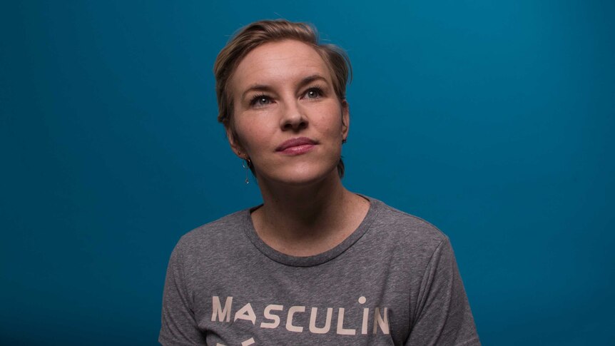 Kate Mulvany, wearing a grey T-shirt, sits in front of a blue backdrop and looks up.