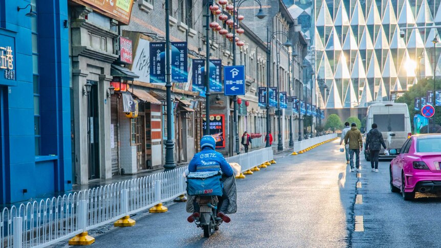 A person rides on a motorbike and people in jackets walk along a street with sunlight reflecting off a building in the distance.