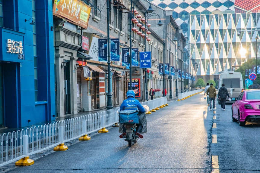 A person rides on a motorbike and people in jackets walk along a street with sunlight reflecting off a building in the distance.