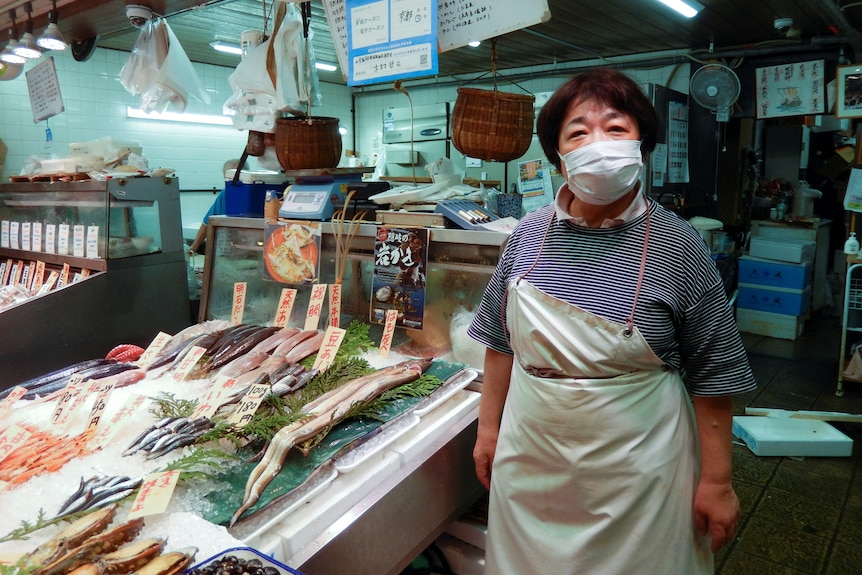 A masked middle-aged Asian woman  wearing an apron stands next to fish cuts on display