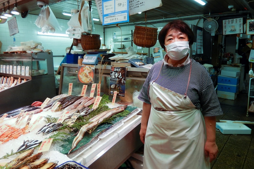 Una mujer asiática de mediana edad enmascarada que usa un delantal se encuentra junto a los cortes de pescado en exhibición