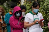 People in face masks stand in line on the street for a COVID-19 test. Many are looking at their phones.