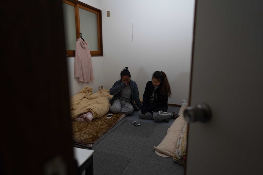 Two women wearing activewear sit on the flood in darkened room with makeshift beds to their left and right.