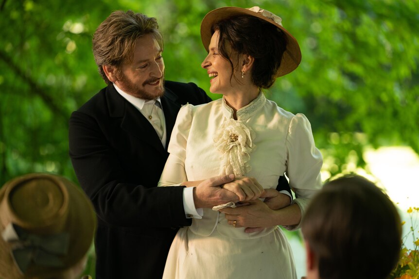 A film still of Benoît Magimel with his arms around a laughing Juliette Binoche. They're both in late 19th century dress.