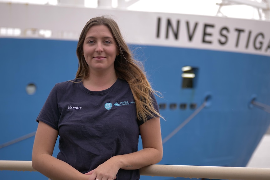 A young woman with brown hair stands in front of a ship named Investigator