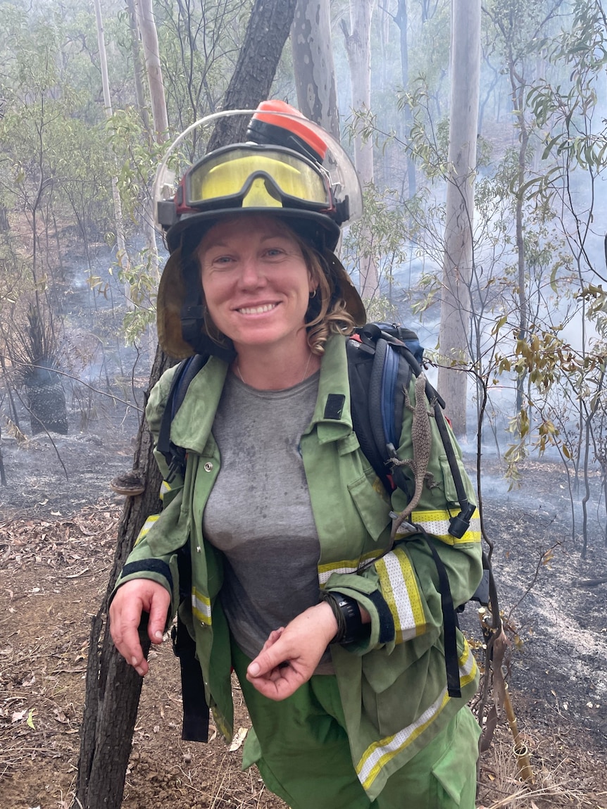 Woman in fire fighting uniform standing in front of smoking bush, smiling