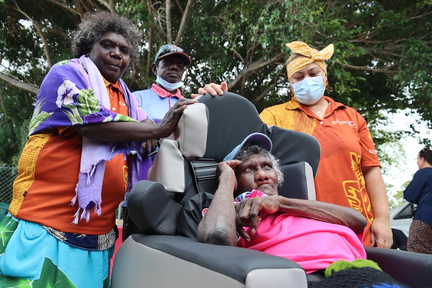 Three people gathered around a woman sitting in a wheelchair, under a large tree. 