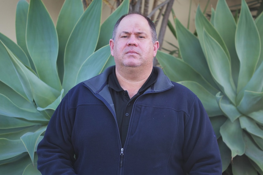 An unsmiling heavyset man, balding, wears a blue fleece jacket and a black tee, stands in front of plants.