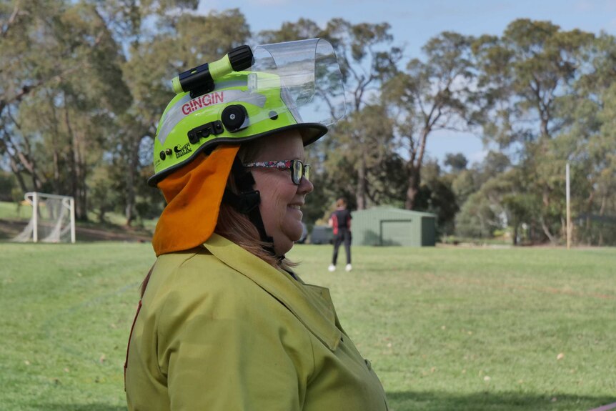 Side profile of Nikki Wood wearing a yellow fire fighting jacket and a fire fighting helmet with visor. Standing on school oval.