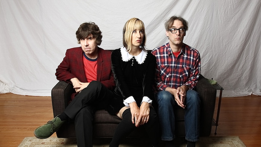 Three members of The Muffs sitting on a couch in a studio with a sheet behind them, not looking at the camera.