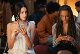 Still from tv episode. Two well-dressed teen girls sit next to each other, looking at their phones.