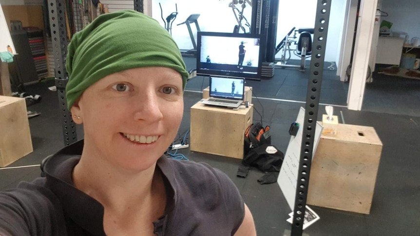 Lady with headscarf stands in front of a home gym setup, with computer screens showing her image.