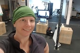 Lady with headscarf stands in front of a home gym setup, with computer screens showing her image.