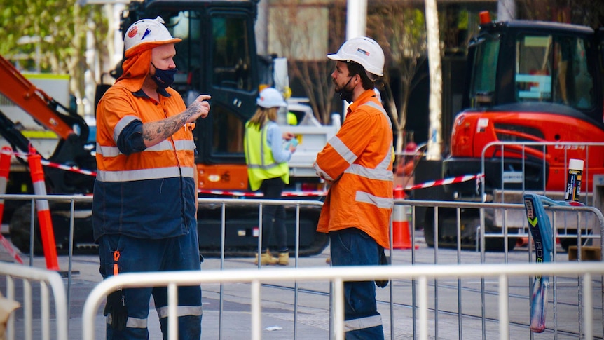 Two construction workers wearing masks standing at a roadworks site.