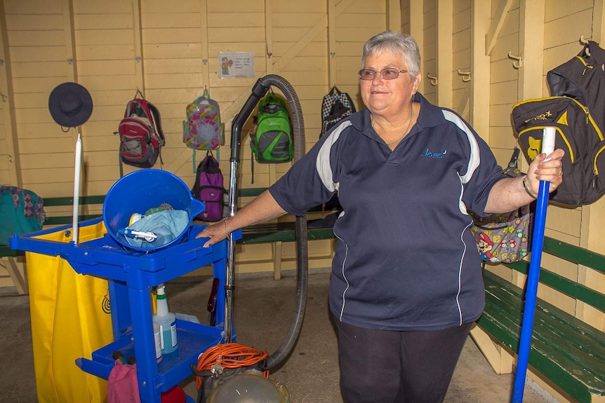 A woman holding a broom in one hand and with her other on a cleaning cart in a school cloakroom