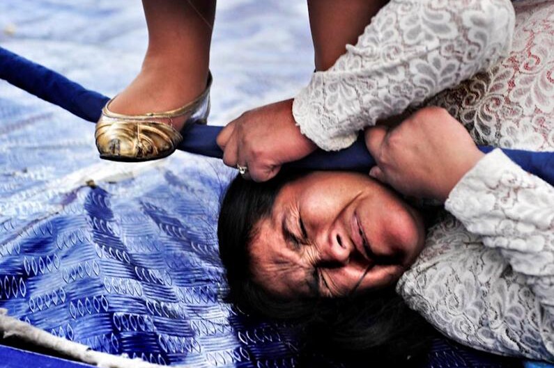 A close-up shows a woman wincing as her opponent stands on her head during a Bolivian wrestling match.