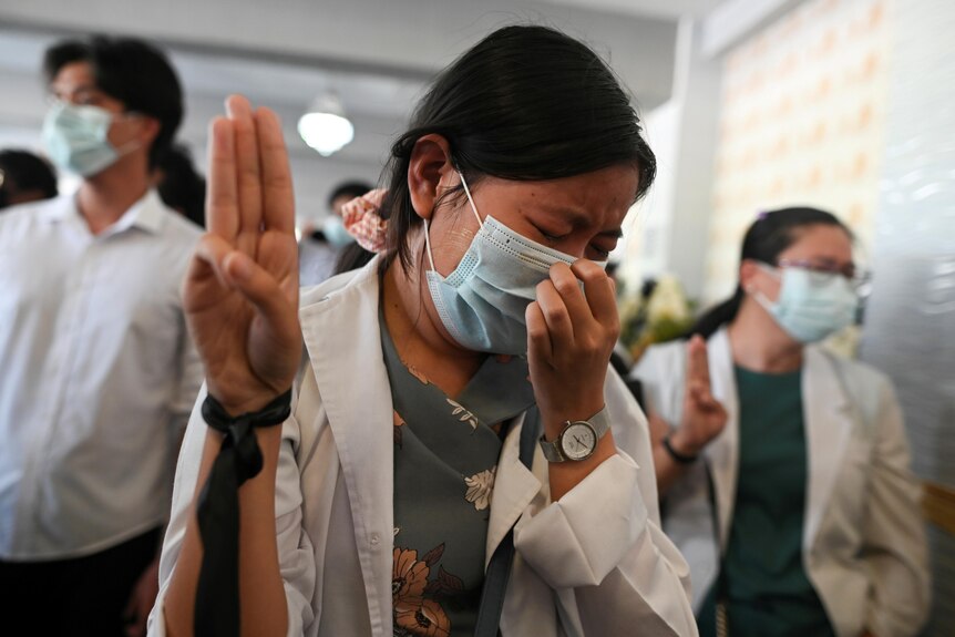 A grieving woman in a white coat and face mask flashes a three-finger salute at funeral as she looks away