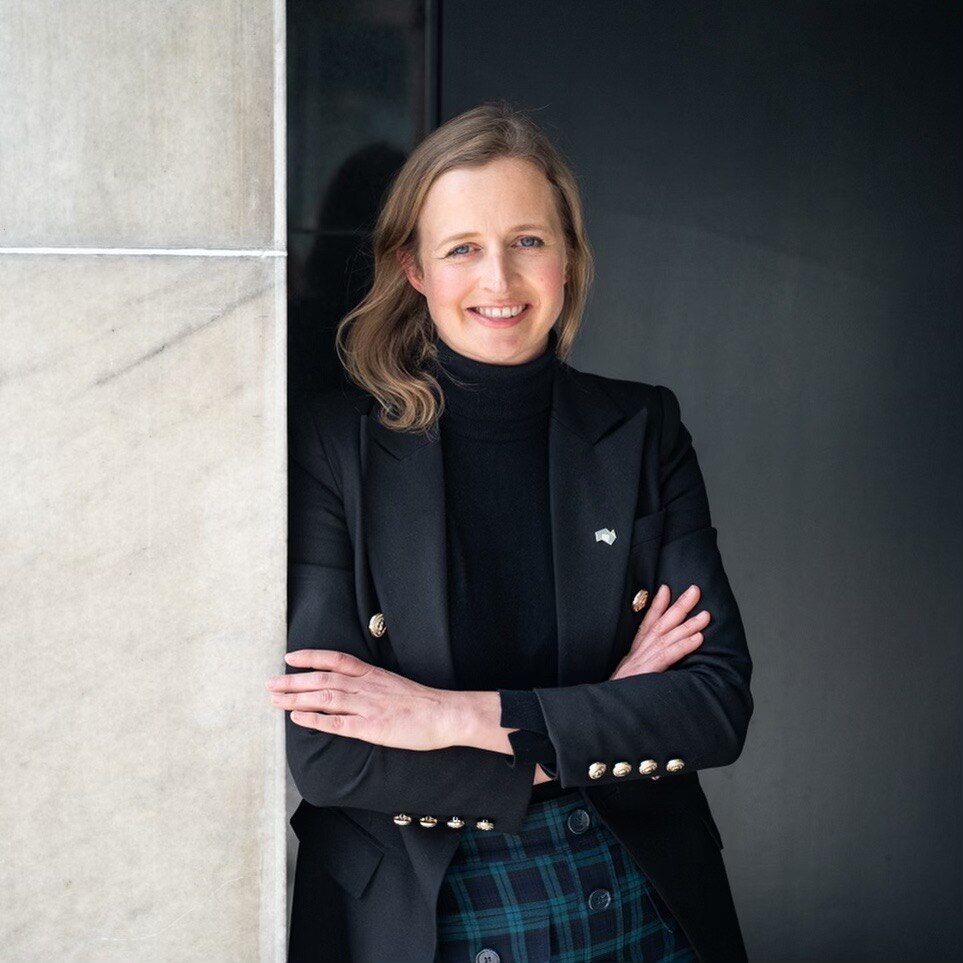 Emily Bourke, standing against a white and black wall with a big smile.