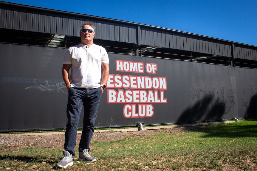 A middle aged man in white t-shirt and jeans stands in front of building which says 'home of Essendon baseball club'