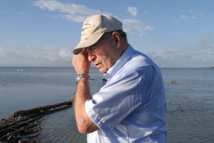 A man in a blue shirt and a baseball cap wipes away a tear with the ocean in the background