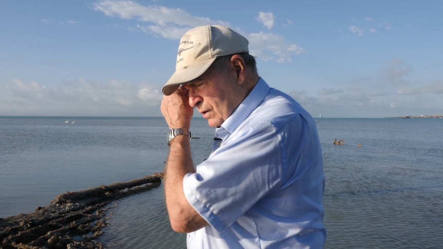 A man in a blue shirt and a baseball cap wipes away a tear with the ocean in the background