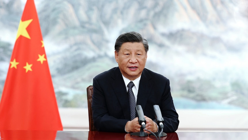 A man wearing a suit and tie sits at a desk in front of a Chinese flag