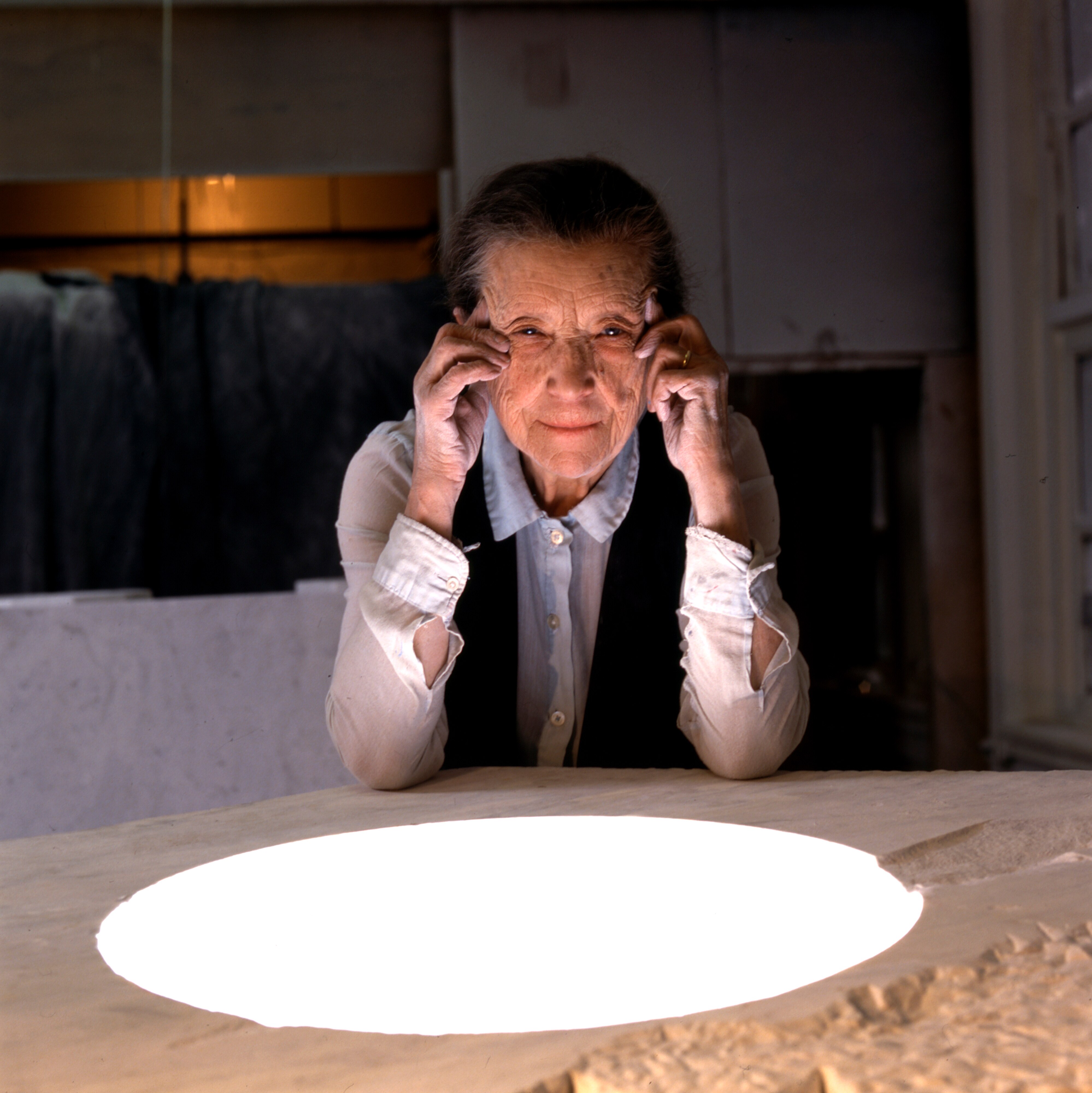 Louise Bourgeois, an elderly white woman wearing a white blouse and black vest sits at an art studio table with head in hands.