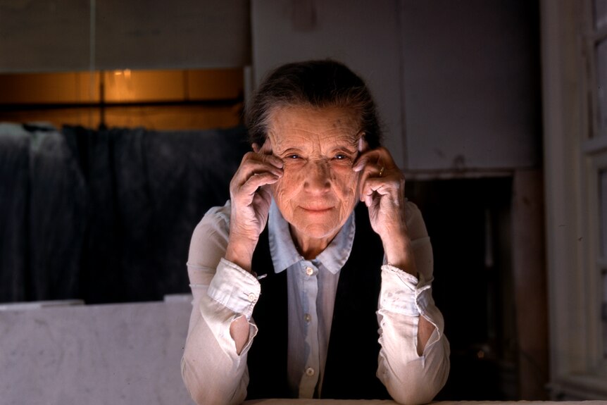 Louise Bourgeois, an elderly white woman wearing a white blouse and black vest sits at an art studio table with head in hands.