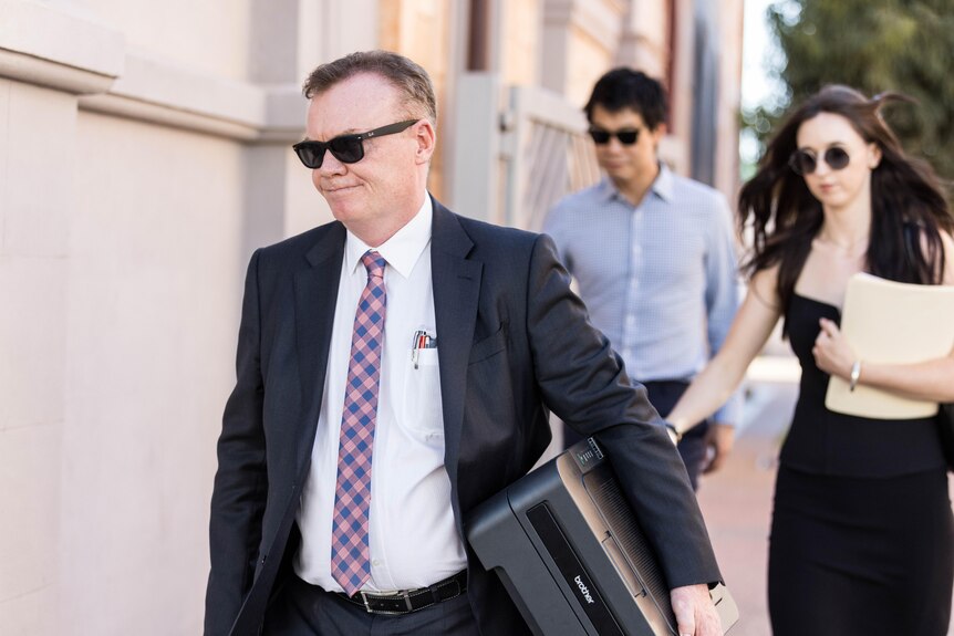 A man in a dark suit and dark sunglasses walks away from a court building.