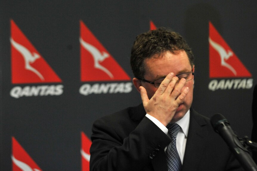 A middle-aged man in a suit with curly hair and glasses touches his face in front of a row of Qantas logos.